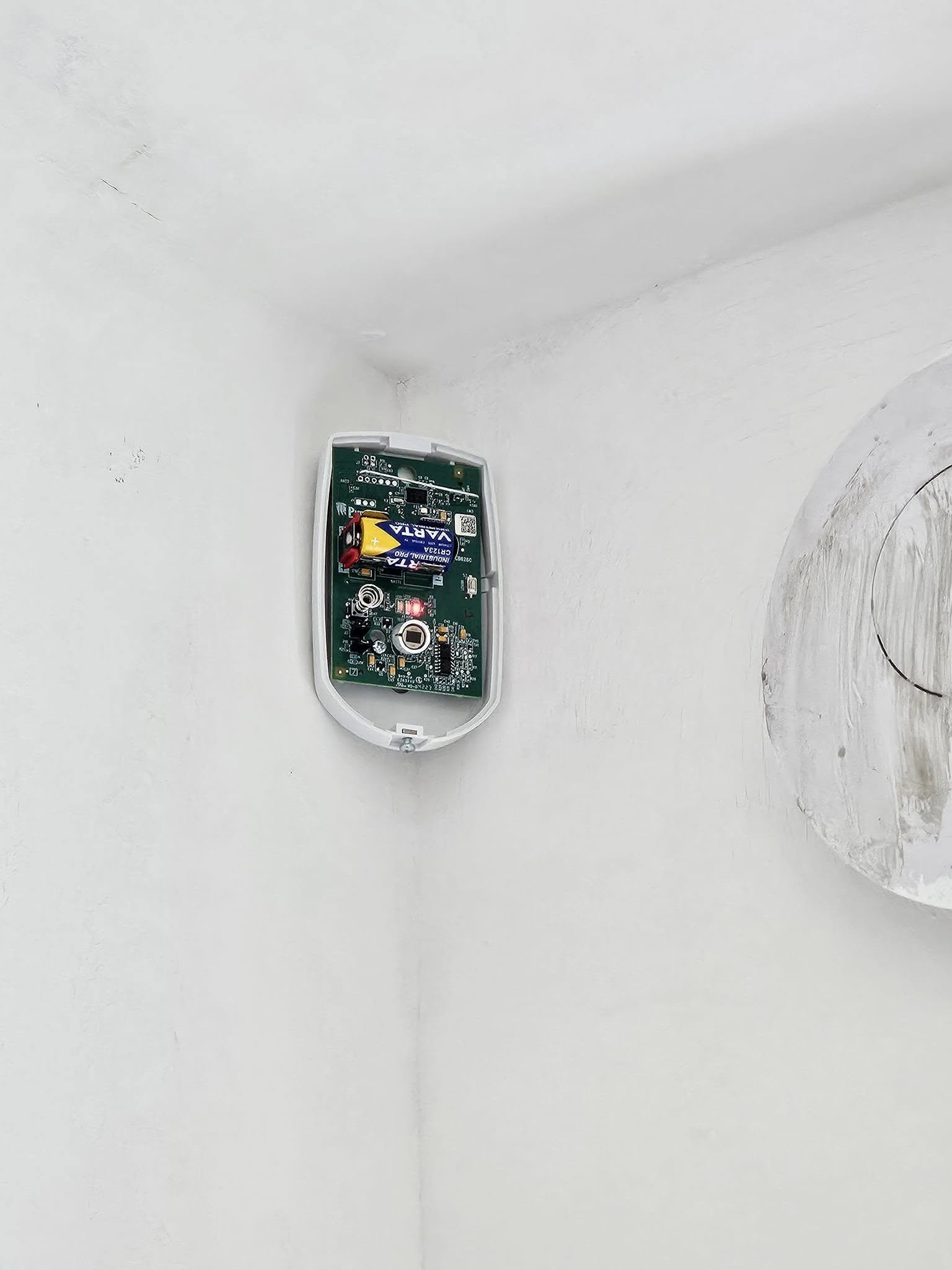 A wireless PIR sensor open on a wall, showing the circuit board and battery, being fitted in a room corner