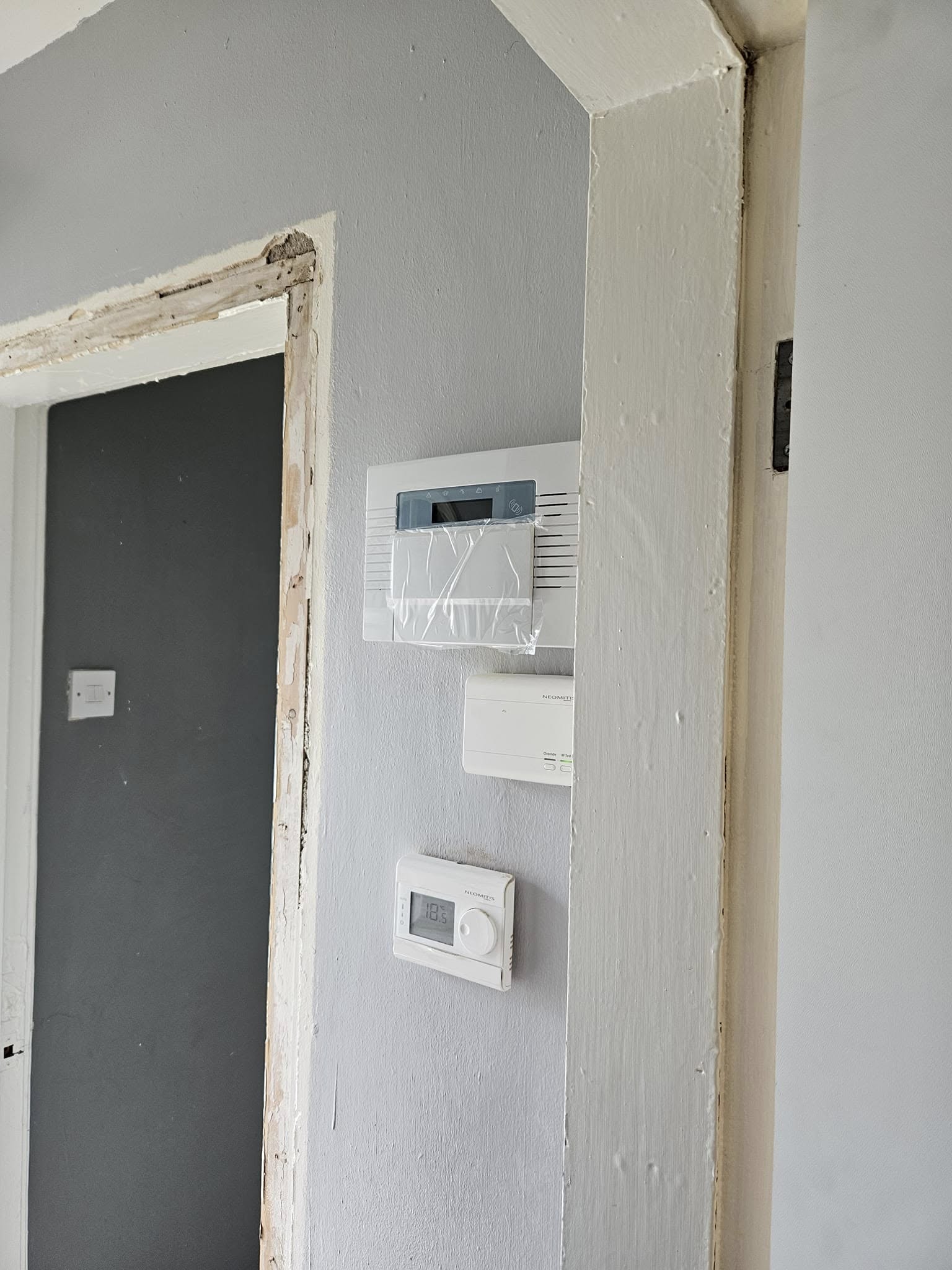 A newly fitted alarm keypad still wrapped in protective film, with a wireless receiver and thermostat below it